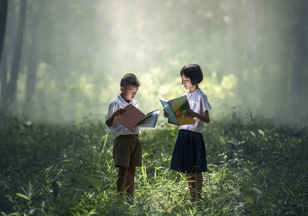 A boy and a girl stand in the grass and read books.