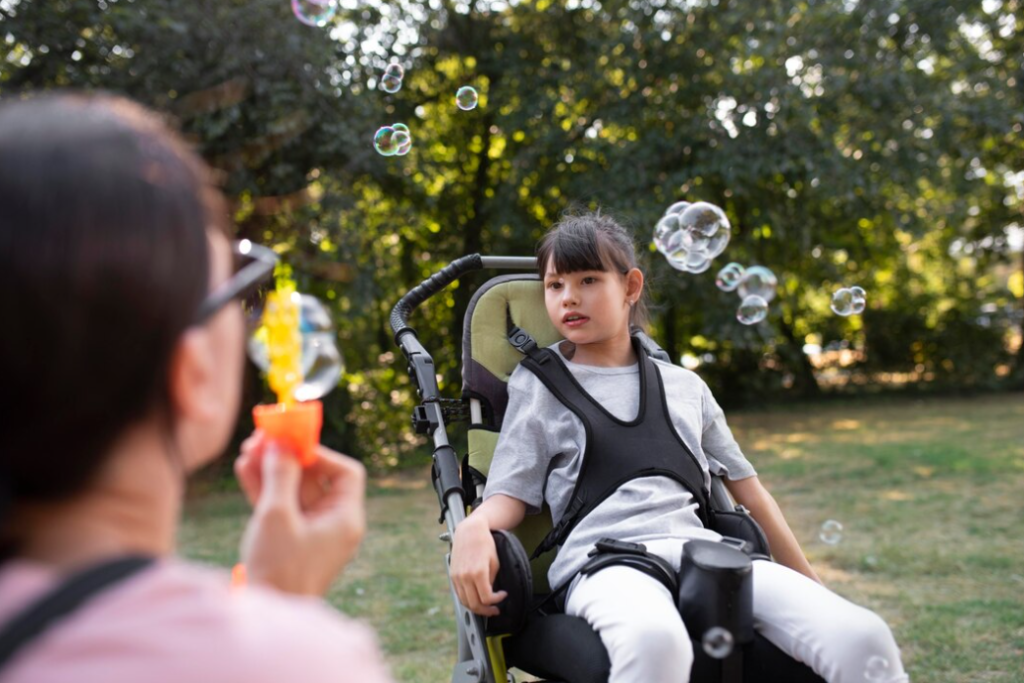A girl in a wheelchair watches bubbles floating around her while an adult blows them in a park.