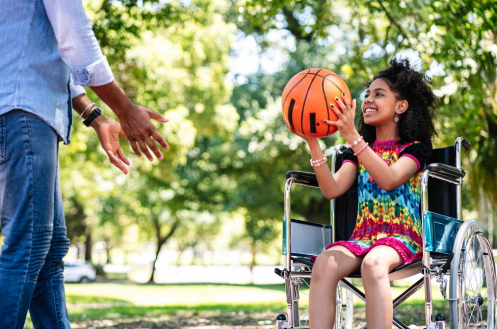 A girl in a wheelchair catches a basketball passed to her in an outdoor setting.