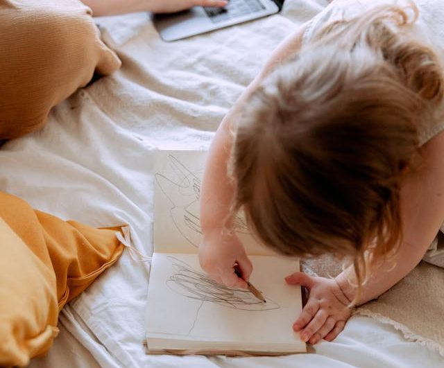 A young girl sitting on a bed, focused on drawing in a book.